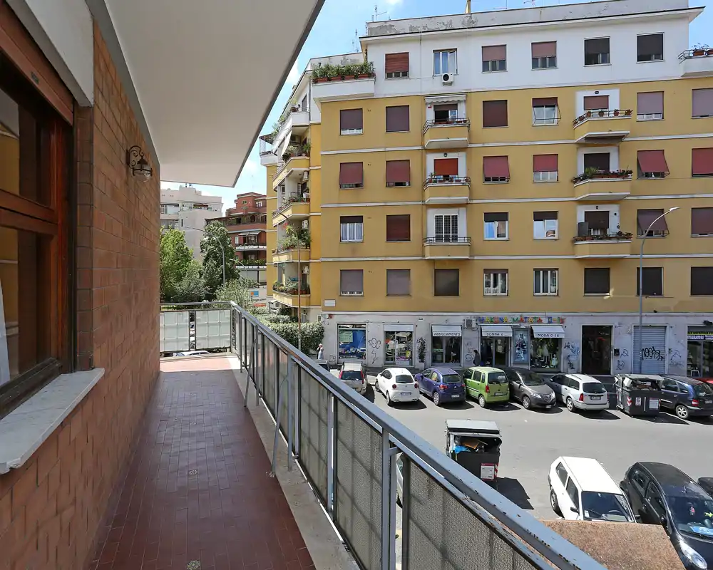 Balcón soleado con vistas a una calle de la ciudad, suelo de baldosas, barandilla metálica y vista de los edificios vecinos—ideal para el café matutino.