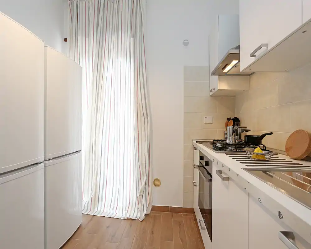 Cocina moderna y compacta con muebles blancos, placa de gas, horno empotrado y frigorífico junto a una ventana iluminada con cortinas a rayas.