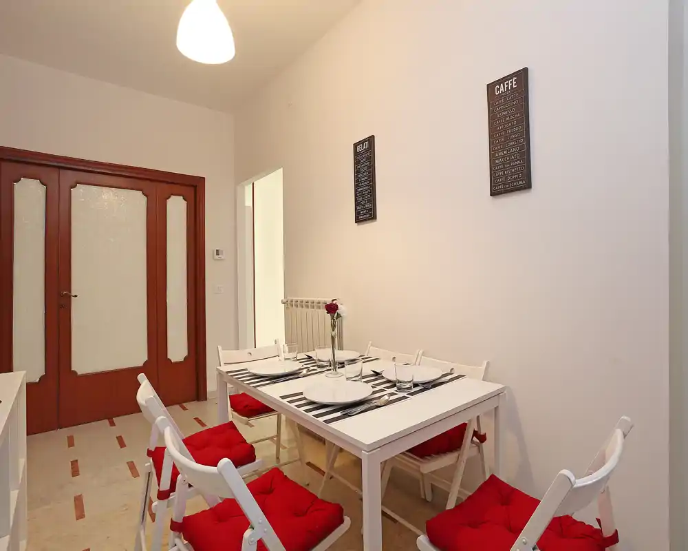 Zona de comedor luminosa y moderna con mesa blanca puesta para cuatro y sillas plegables con cojines rojos, decoración de pared sencilla y una flor como centro de mesa.