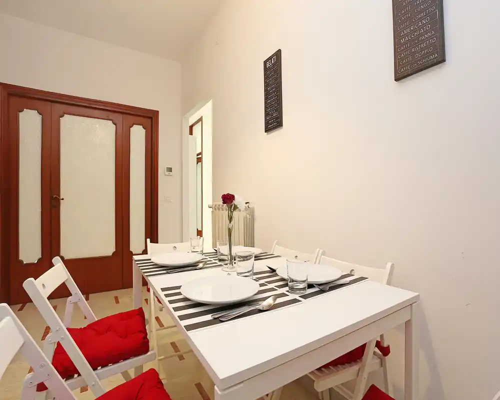 Área de comedor luminosa y ordenada con mesa blanca, manteles a rayas y sillas con cojines rojos; estilo contemporáneo y sencillo ideal para un piso pequeño.