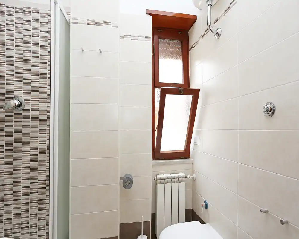Baño compacto y bien iluminado con paredes alicatadas, mampara de ducha a la izquierda, ventana inclinada con marco de madera y radiador; decoración neutra y limpia, ideal para anuncios.