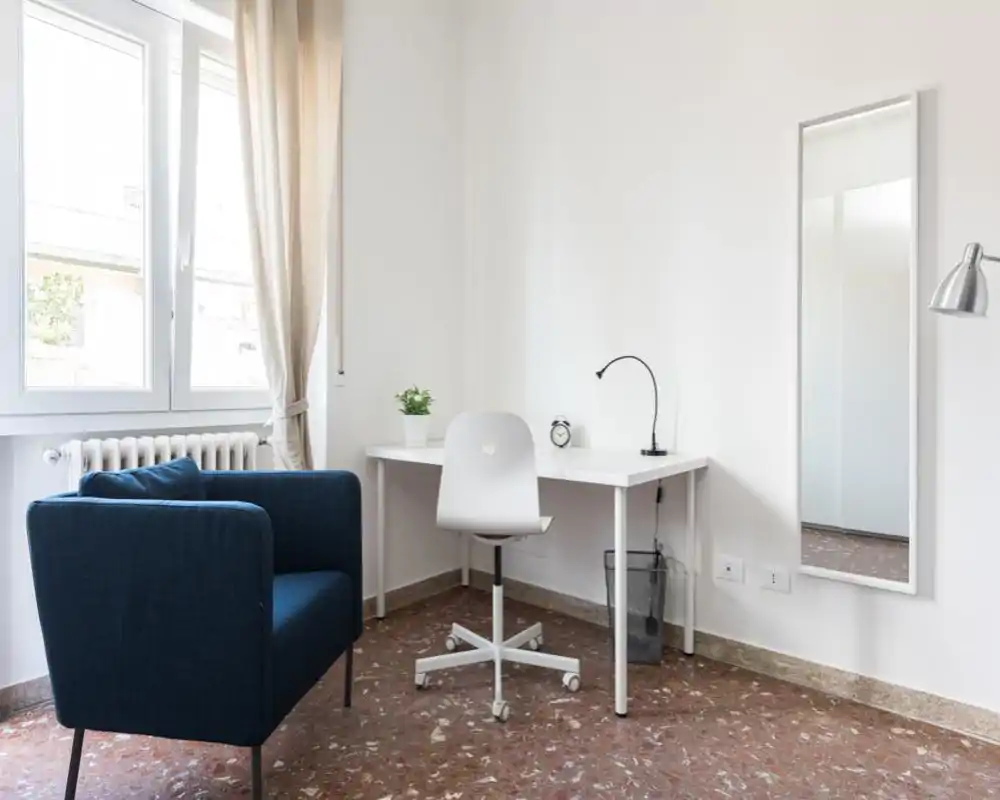 Angolo soggiorno luminoso e minimalista con poltrona blu, scrivania bianca e ampia finestra che offre molta luce naturale — perfetto per relax o lavoro.
