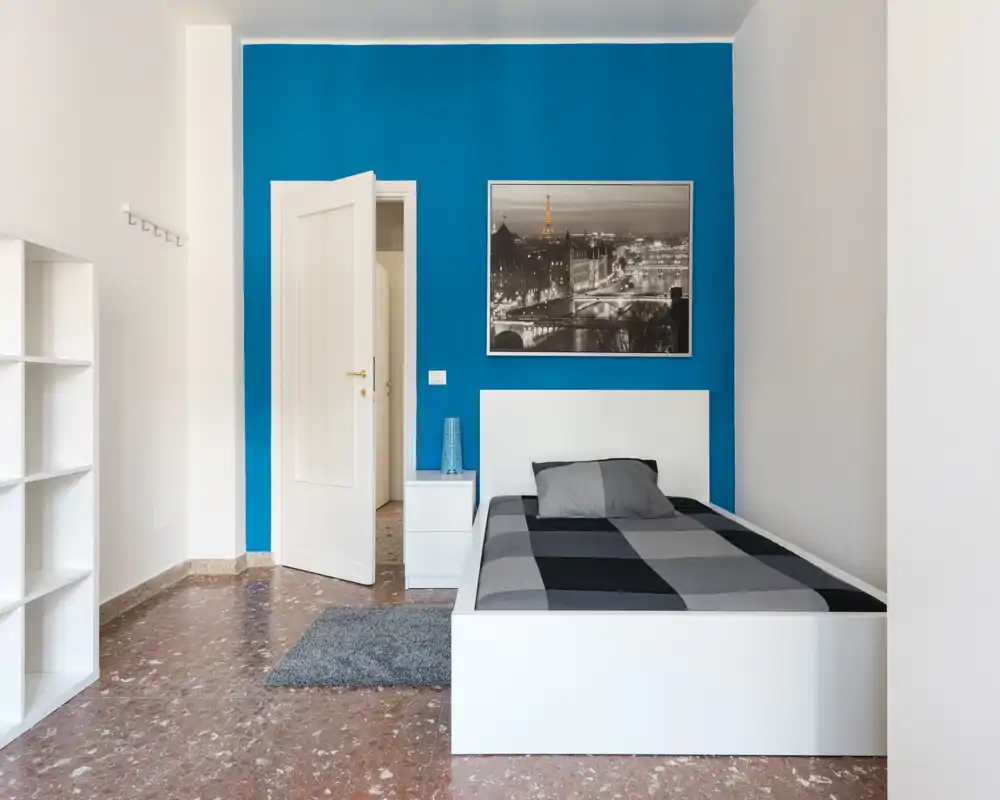 Camera da letto singola luminosa e moderna con struttura del letto bianca su una parete blu vivace, comodino minimal e una foto incorniciata della città.
