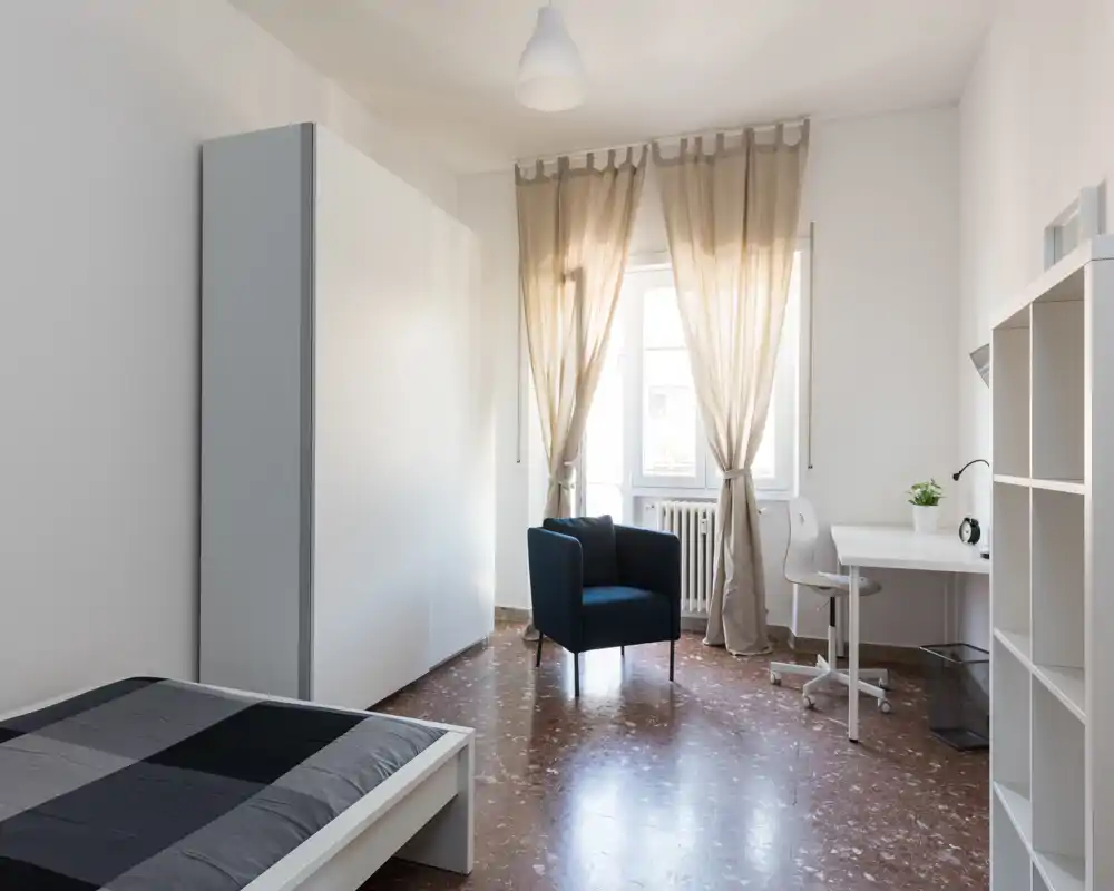 Camera da letto luminosa e minimale con grande letto, armadio, scrivania e una poltrona accogliente vicino alla finestra; luce naturale e arredi neutri creano un ambiente rilassante.