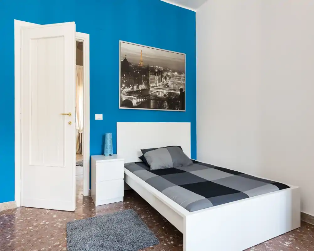 Camera da letto singola, minimal e ordinata con letto bianco basso su una parete accentuata blu, comodino e quadro con foto della città — luminosa e perfetta per soggiorni brevi.