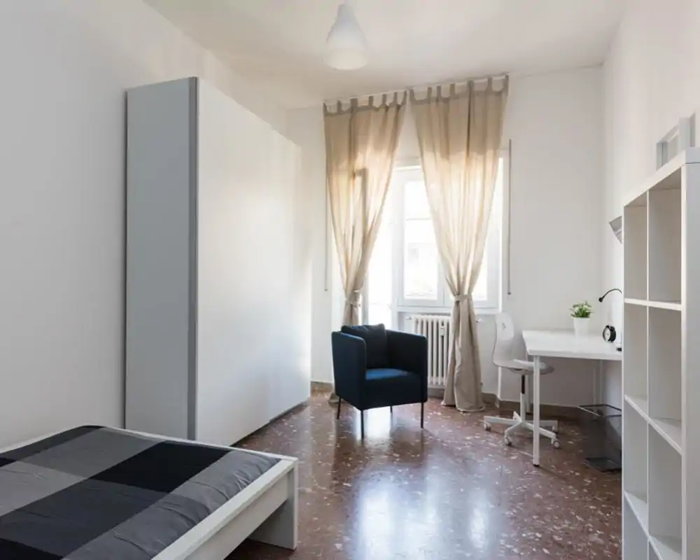Camera da letto luminosa e minimal con letto visibile in primo piano, una poltrona blu vicino alla finestra, scrivania con sedia e scaffalatura: ideale per chi cerca uno spazio arioso e ordinato.