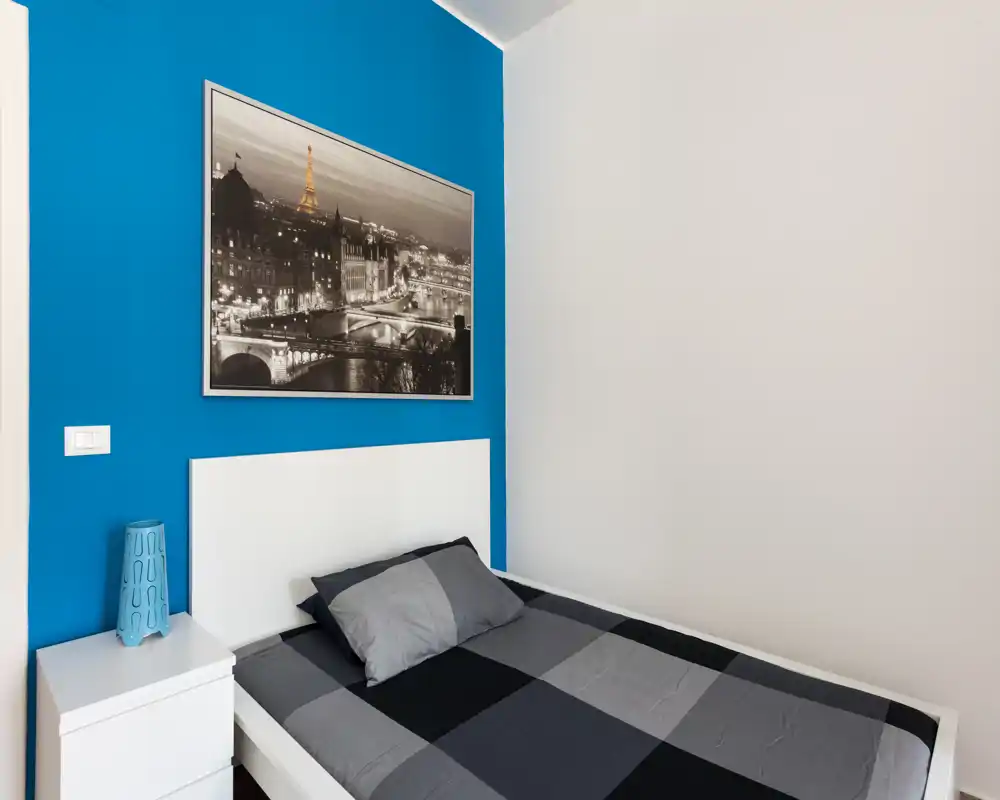 Camera da letto moderna e pulita con struttura del letto bianca, copriletto grigio a quadri e una parete accentuata blu. Comodino semplice con lampada completano l'area notte compatta e luminosa, perfetta per gli ospiti.
