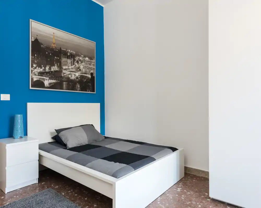 Camera da letto moderna e luminosa con letto bianco su pedana, parete accentuata in blu, biancheria neutra e una stampa incorniciata. Comodino con lampada e un design pulito e accogliente.