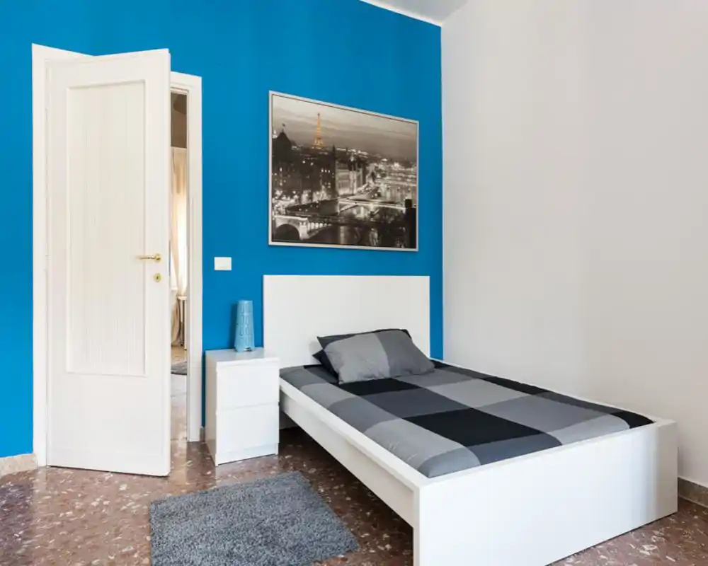 Camera da letto singola, luminosa e minimale con letto bianco, comodino e una parete accentuata blu con quadro appeso.