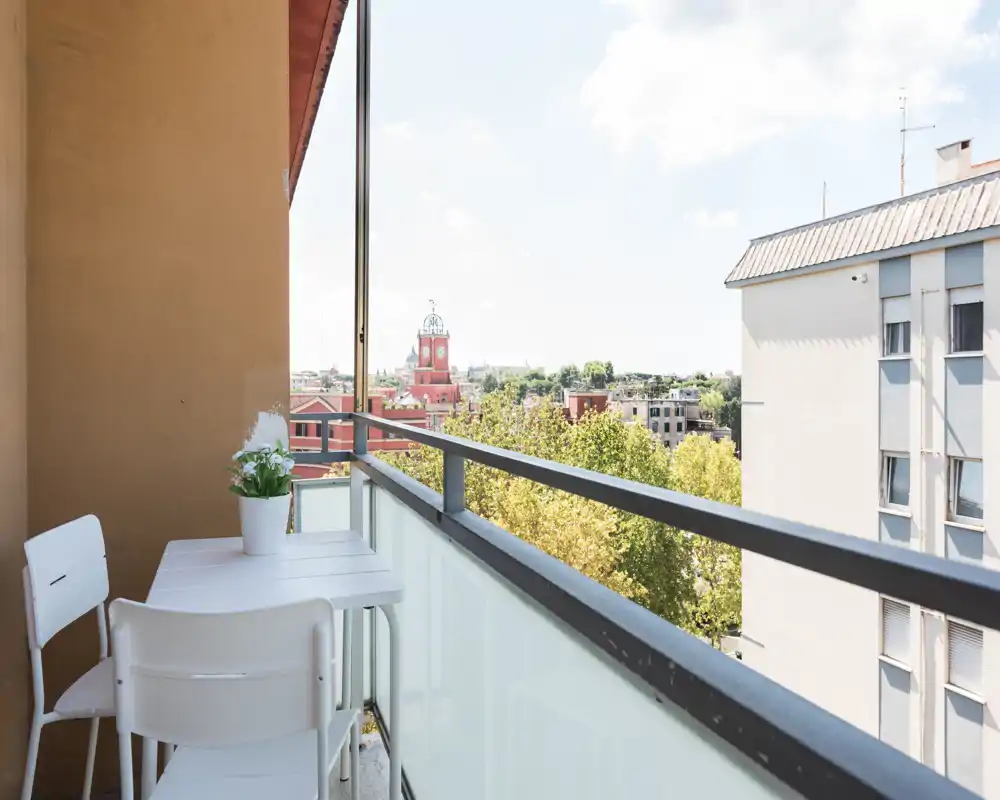 Balcone soleggiato con piccolo tavolo e sedie bianche, vaso di fiori e vista aperta su alberi e tetti della città — perfetto per un caffè mattutino.