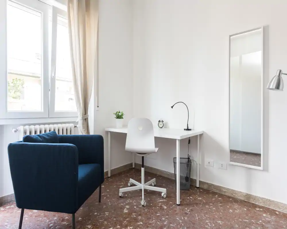 Angolo living luminoso e minimal con poltrona blu scuro, scrivania e sedia bianche vicino a una finestra ampia; stile moderno e semplice, ideale come angolo studio.