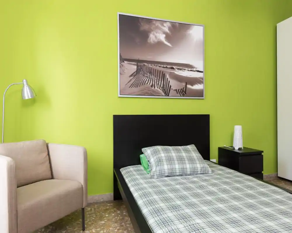 Bright, modern bedroom with a single bed dressed in plaid linens, a black headboard, bedside table and a comfortable armchair—fresh green accent wall and framed seaside artwork complete the look.