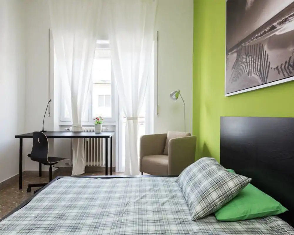 Bright, modern bedroom with a large bed in the foreground, a desk by the window and a comfortable armchair beside a fresh green accent wall.