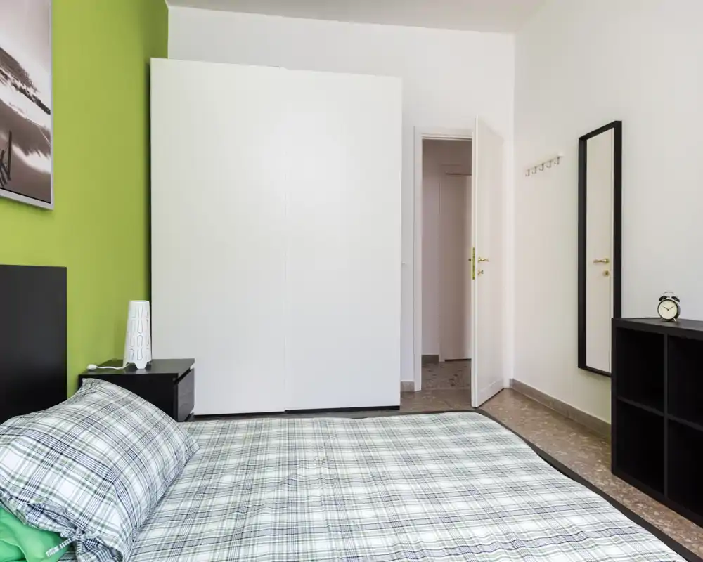 Bright, modern bedroom with a double bed dressed in checked linens, a green accent wall and a large white wardrobe. Clean, minimal furnishings and a full-length mirror create a tidy, welcoming space ideal for rental listings.