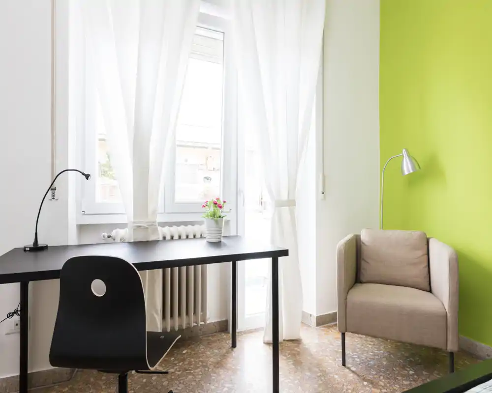 Bright, minimal living area with a desk, chair and a comfortable armchair next to a green accent wall; large window with sheer curtains provides ample natural light.