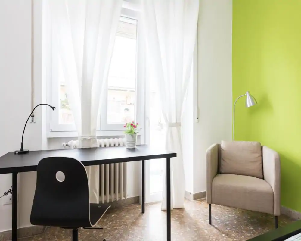 Bright and tidy living area with a desk by a large window, a comfortable armchair and a green accent wall — ideal for a relaxed workspace or reading corner.