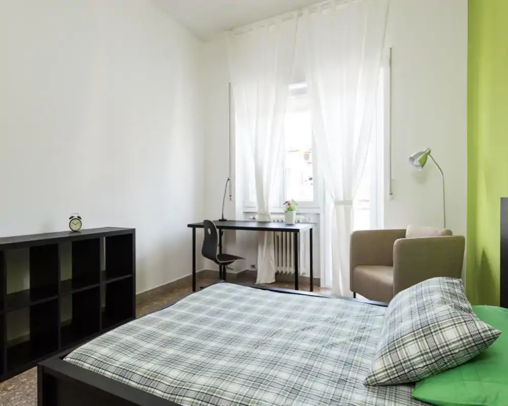 Bright, modern bedroom with a double bed in the foreground, a workspace by the window and a comfy armchair. Clean lines, green accent wall and sheer curtains provide an airy, inviting atmosphere.