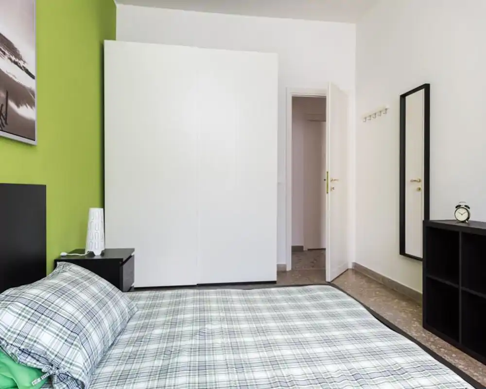 Bright, modern bedroom with a plaid double bed in the foreground, a large white wardrobe against a green-accent wall, bedside table with lamp and a full-length mirror near the door.