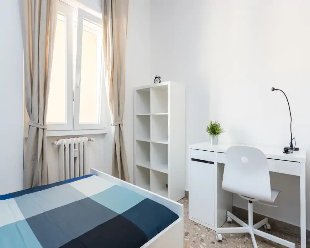Bright, minimal bedroom with a neatly made bed, a study desk and shelving. Natural light from the window creates a clean, airy atmosphere ideal for long-term rentals.