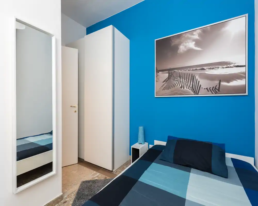 Bright, compact bedroom with a blue accent wall, wardrobe, mirror and a neatly made double bed. Clean, modern styling and good natural/artificial light make this room inviting for short stays.
