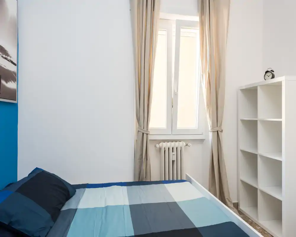 Bright, simply furnished bedroom with a double bed dressed in blue geometric bedding, a window with beige curtains letting in natural light, and a white shelving unit for storage.