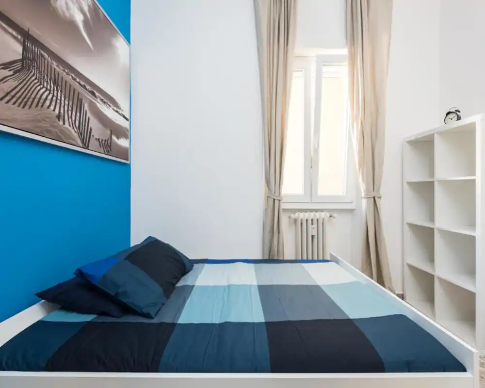 Bright, modern bedroom with a blue accent wall and neatly made double bed. Neutral curtains and shelving offer practical storage and a calm atmosphere.