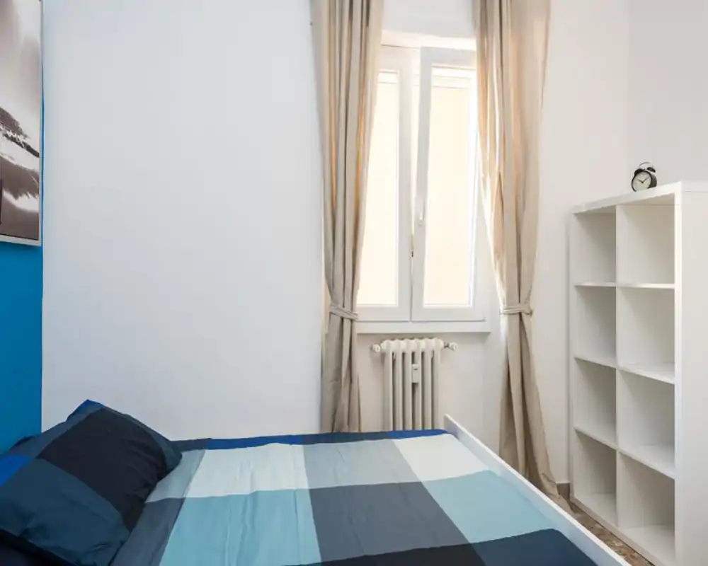 Bright, simply furnished bedroom with a double bed dressed in blue linens, a window with beige curtains letting in natural light, and a white shelving unit for storage.