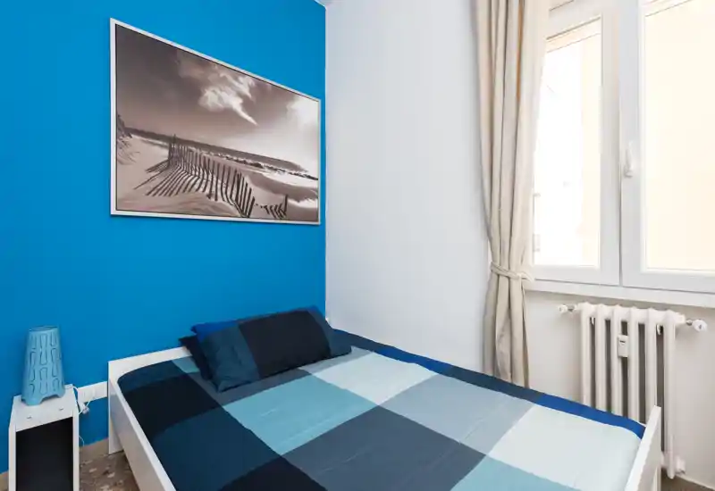 Compact, modern bedroom with a double bed against a vivid blue accent wall, coordinated blue bedding, framed coastal artwork and natural light from a window.