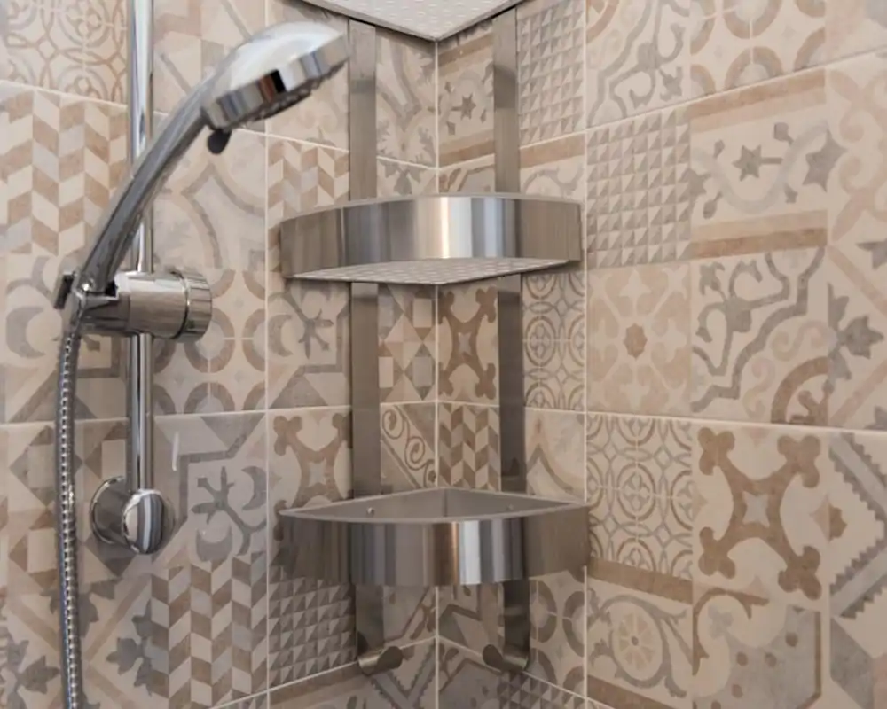 Close-up of a modern shower corner with chrome handheld shower head and stainless-steel corner shelf against patterned ceramic tiles.