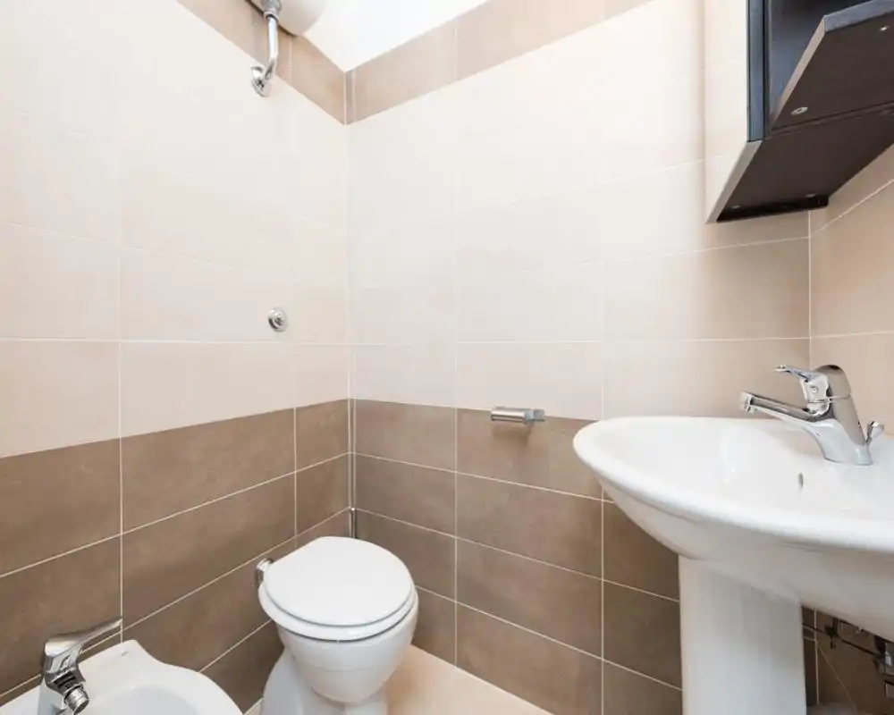 Clean, compact bathroom with white sink, toilet and bidet set against neutral beige wall tiles; bright, well-lit and freshly maintained.