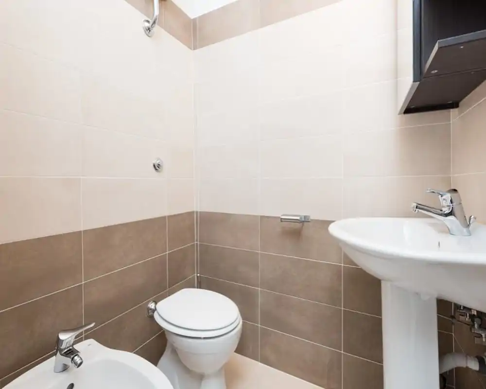 Clean, modern bathroom with a toilet, bidet and pedestal sink set against two-tone beige wall tiles. Bright, minimalist finish ideal for a rental listing focused on hygiene and simplicity.