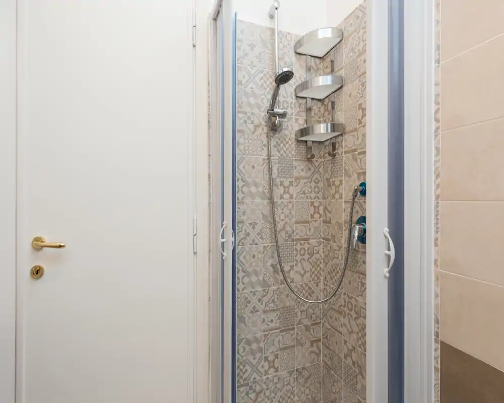 Compact bathroom shower with patterned ceramic tiles, a handheld showerhead and metal corner shelves — clean, modern and well lit.