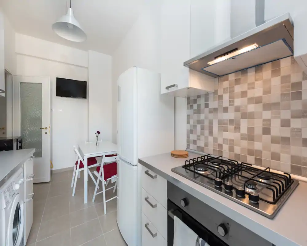 Bright modern kitchen with a gas hob, oven and fridge, plus a small dining table with chairs — clean, compact and well lit.