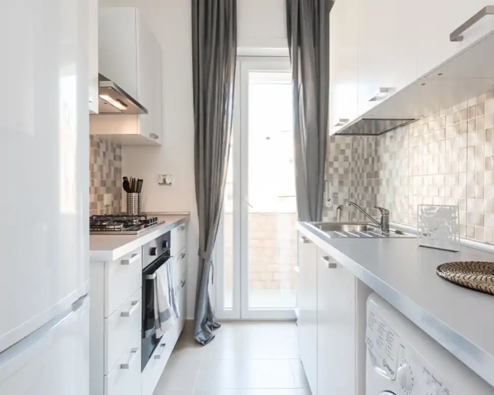 Bright modern galley kitchen with white cabinets, tiled backsplash, gas stove and a sink by a glass door letting in natural light.