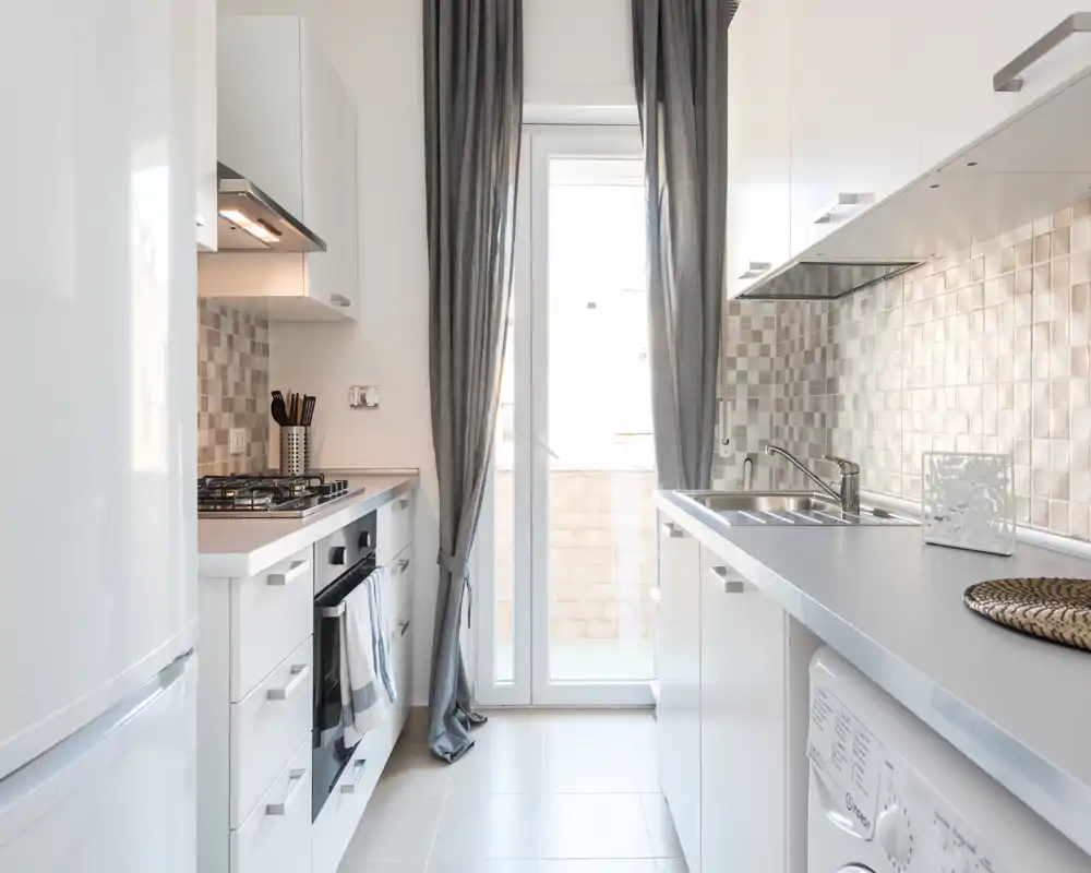 Bright modern galley kitchen with white cabinetry, built-in oven and gas hob, stainless sink and a glass door leading to a small balcony — neat, well-lit and functional.