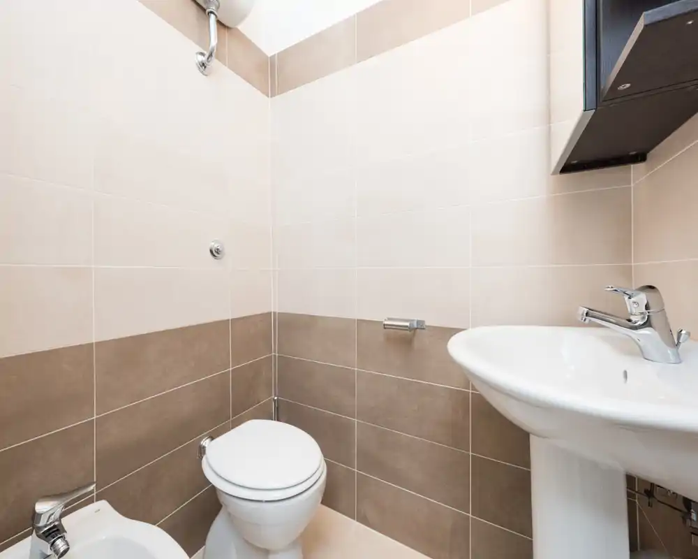 Compact, well-lit bathroom with pedestal sink, toilet and bidet against beige and brown tiled walls — clean and functional.