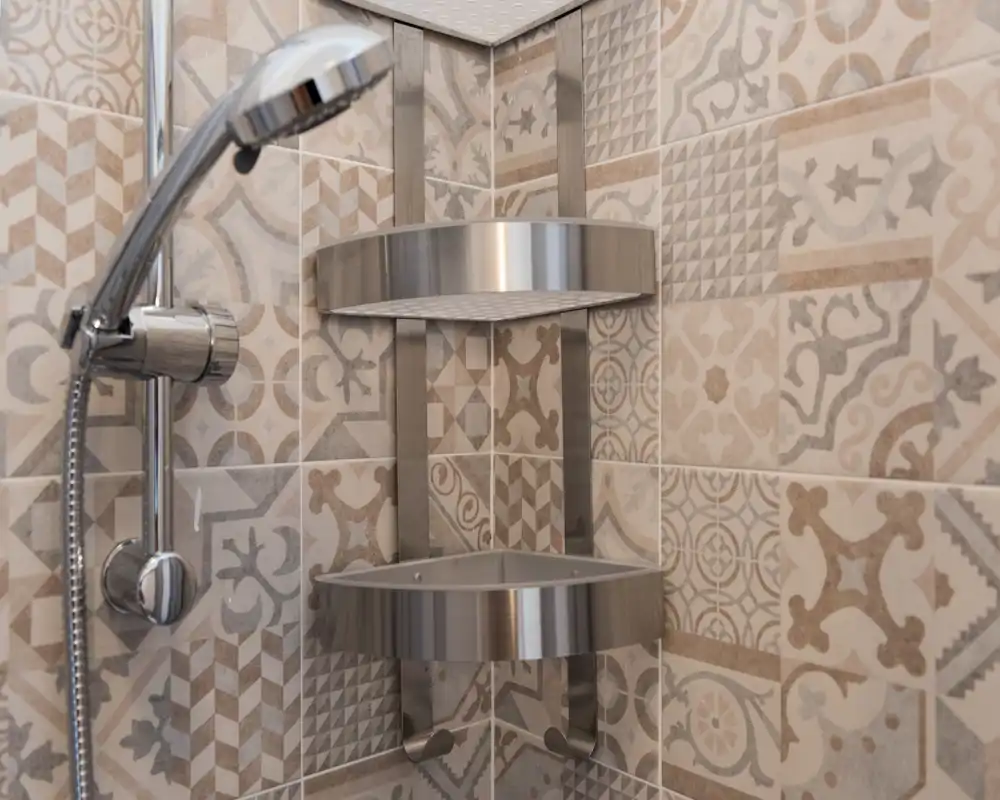 Close-up of a modern shower corner with patterned ceramic tiles, a chrome handheld showerhead and a stainless-steel corner shelf.