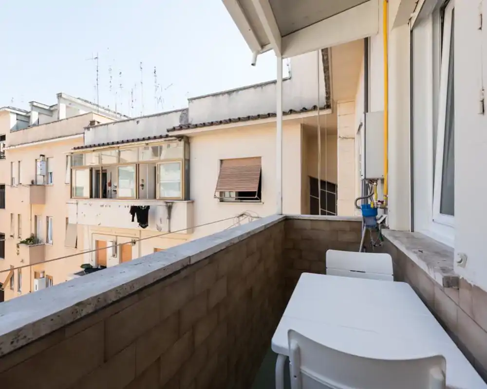 Small private balcony with a compact table and chairs, overlooking neighboring buildings. Bright daylight and simple urban view make it suitable for morning coffee or drying clothes.