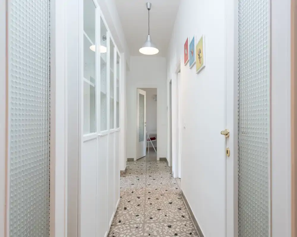 Bright, narrow entrance corridor with patterned tile floor, white walls and built-in storage; well-lit by a central pendant lamp and natural light from the far room.