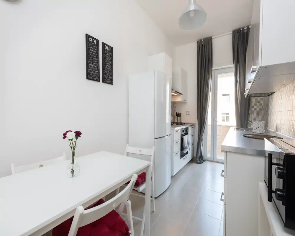 Bright, modern galley kitchen with white cabinetry, integrated oven and fridge, small dining table and natural light from a balcony door — ideal for a compact apartment.