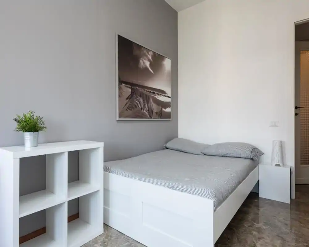 Camera da letto moderna e minimale con letto bianco con biancheria grigia a righe, comodino e quadro decorativo; luminosa, ordinata e adatta a foto per affitti.