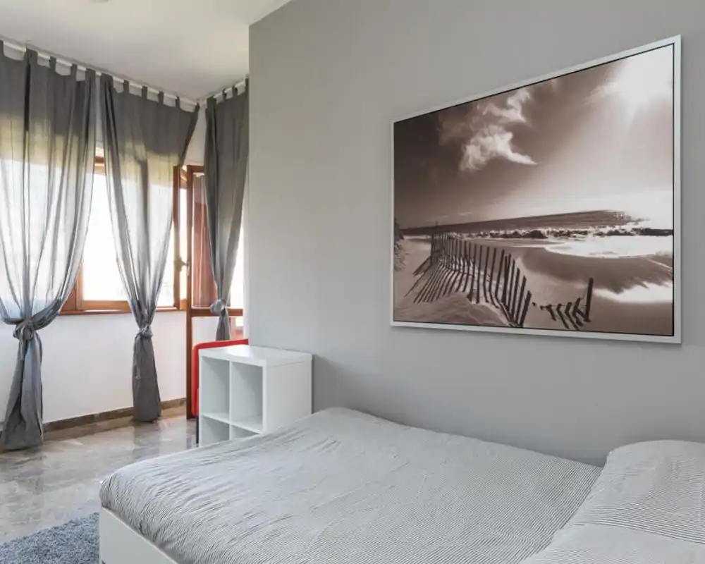 Camera da letto luminosa e minimalista con letto matrimoniale ordinato, ampia finestra con tende annodate e grande quadro marino sopra il letto—palette neutra e pavimento in marmo.