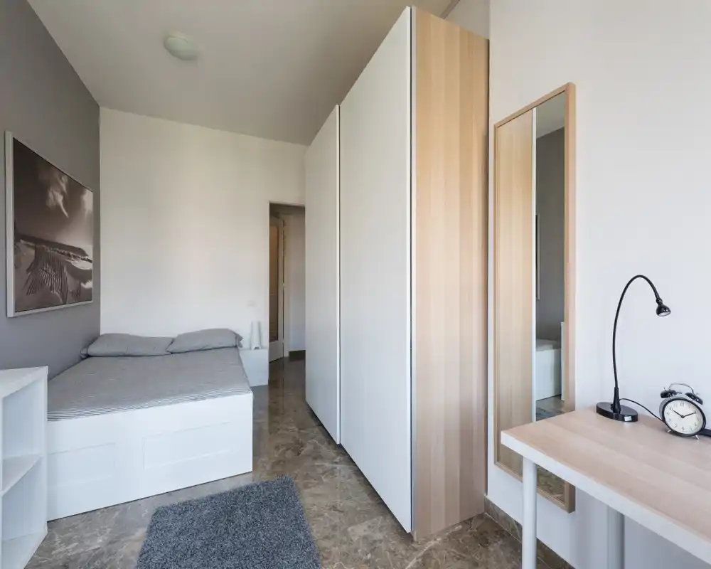 Camera da letto moderna e luminosa con un letto su pedana bianco, grande armadio e una scrivania minimalista. Toni neutri e pavimento in marmo creano un’atmosfera tranquilla e ariosa, perfetta per annunci.