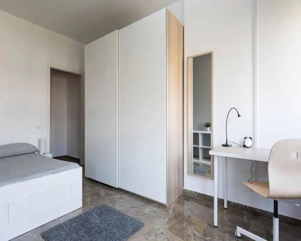 Camera da letto minimalista con letto bianco, ampia armadiatura e una scrivania compatta. Spazio luminoso e ordinato con toni neutri e un piccolo tappeto che aggiunge profondità.