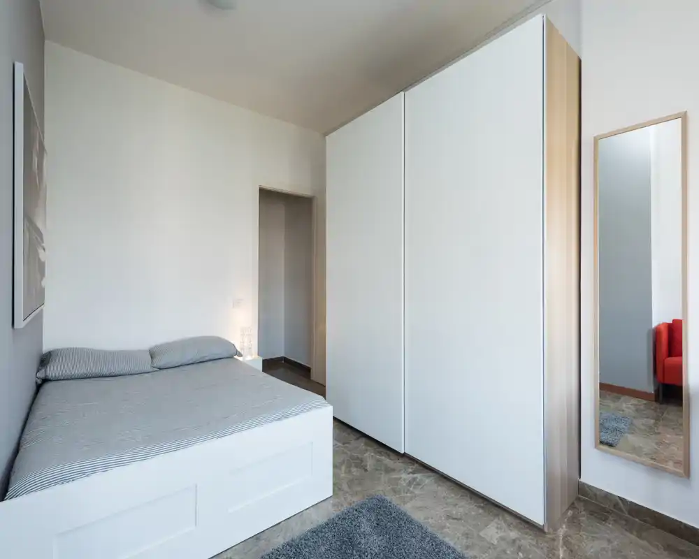 Camera da letto moderna e semplice con letto matrimoniale, grande armadio bianco e specchio intero; luce naturale intensa e arredo minimo e ordinato.