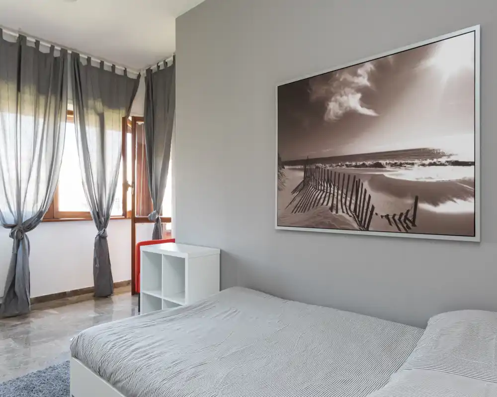 Camera da letto luminosa e minimal con un grande letto in primo piano, biancheria neutra, luce naturale dalle finestre e una stampa marina incorniciata sulla parete.