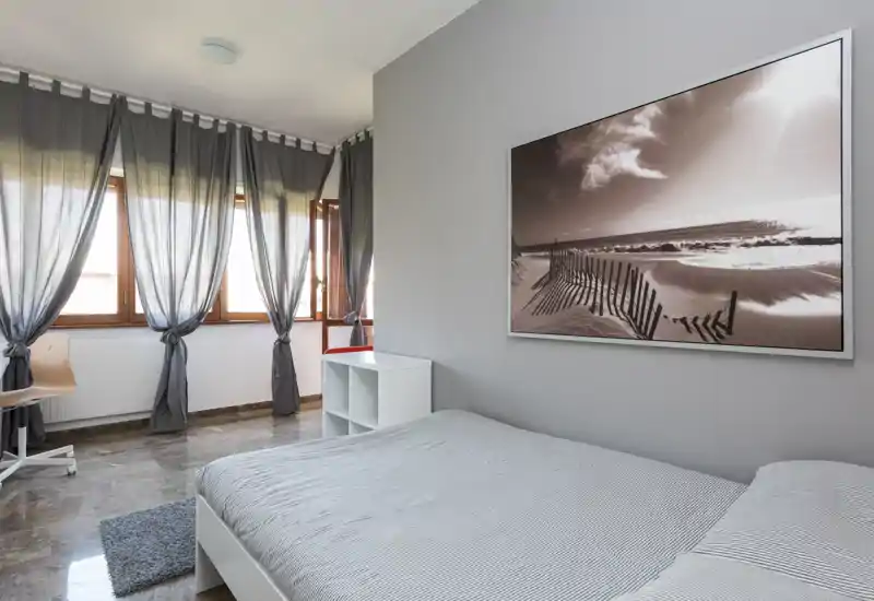Camera da letto luminosa e minimale con letto matrimoniale, ampie finestre con tende leggere, piccolo mobile a cubi e quadro a parete. La luce naturale riempie lo spazio conferendo un’atmosfera calma e ariosa, adatta per gli annunci.