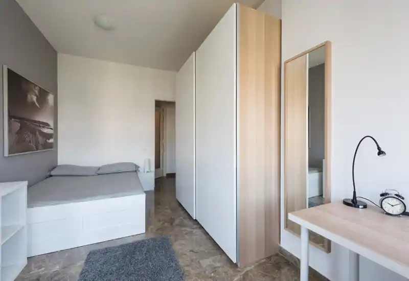 Camera da letto moderna e luminosa con un letto su pedana bianco, grande armadio e una scrivania minimalista. Toni neutri e pavimento in marmo creano un’atmosfera tranquilla e ariosa, perfetta per annunci.