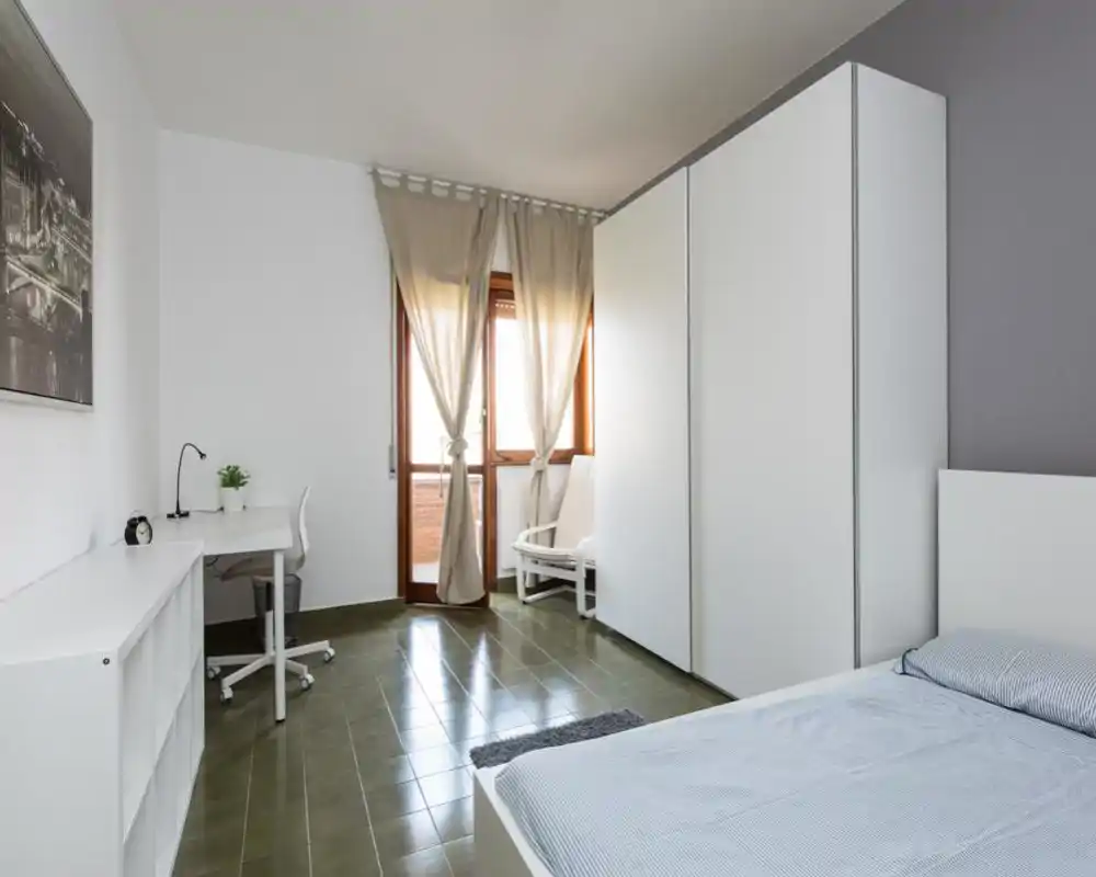 Camera da letto luminosa e minimale con letto matrimoniale visibile a destra, ampio armadio bianco, scrivania e sedia vicino alla finestra e tende leggere che filtrano la luce naturale.