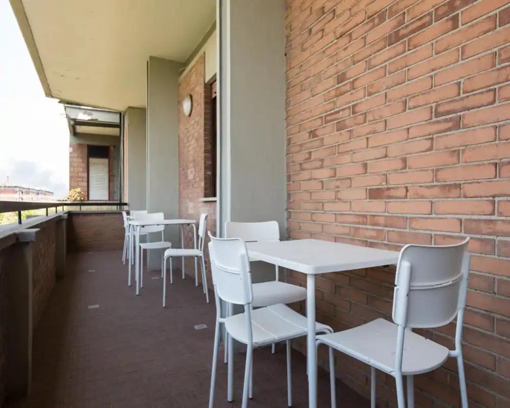Balcone soleggiato arredato con tavolo e sedie bianche da esterno contro una parete in mattoni — ideale per pranzi informali o per rilassarsi all'aperto.